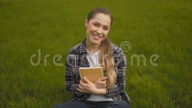 中档。 20岁的女孩穿着格子衬衫坐在草地上，真诚地笑了